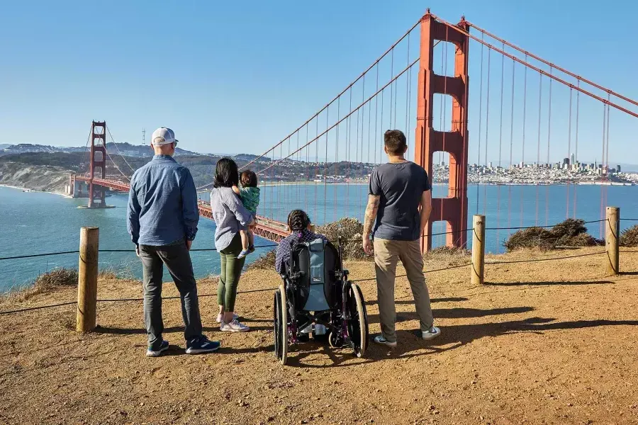 Un gruppo di persone, tra cui una persona in sedia a rotelle, è ripreso da dietro mentre osserva il Golden Gate Bridge da Marin Headlands.